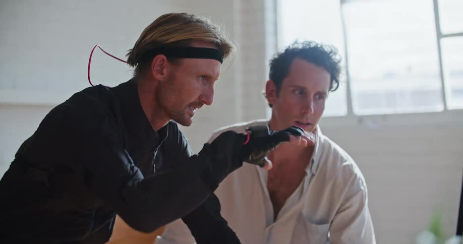 A man with virtual reality equipment on stands in an office next to another man and they look at a computer as they move and close their hands