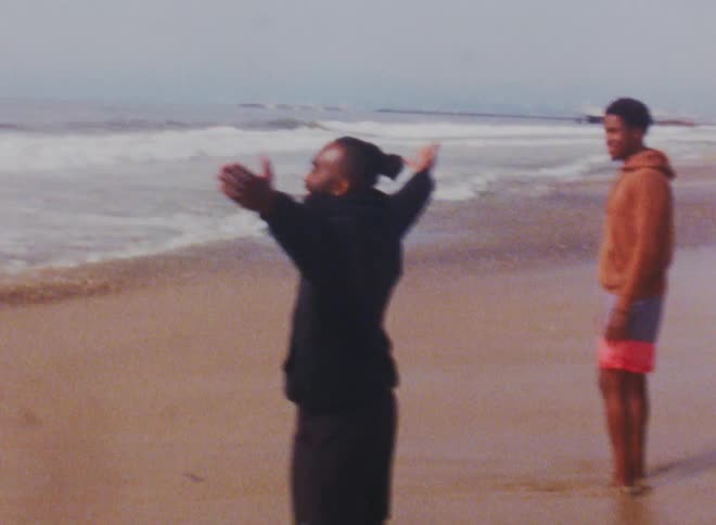 Two men stand on the beach and one man yells and raises his hands to his side