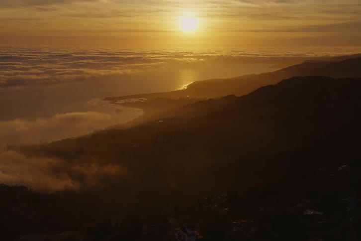 The sun is shining brightly above clouds and fog and reflecting onto the water along the coast next to mountains