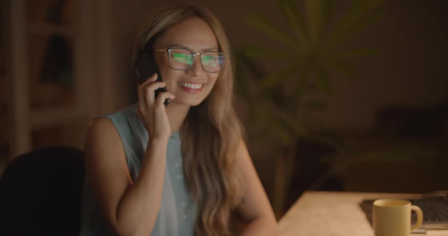 Woman with long brown hair glasses and blue blouse receives call while typing on computer keyboard