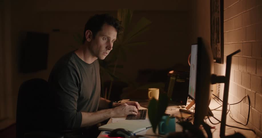 A man in an office holds a book and moves his chair in front of his computer and starts typing