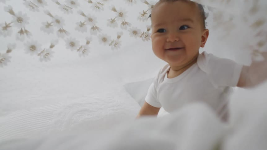 A baby plays under lace fabric while crawling in a bed
