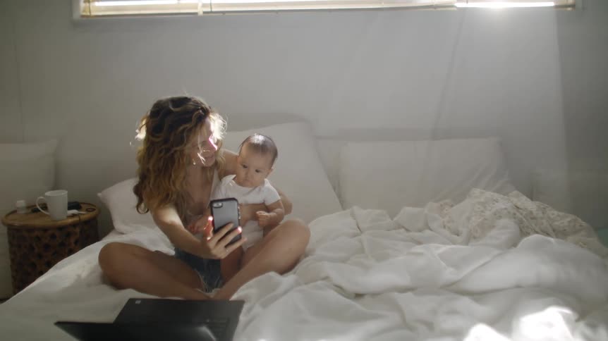 A young woman sits in bed and holds her baby and takes a selfie of them with her cell phone