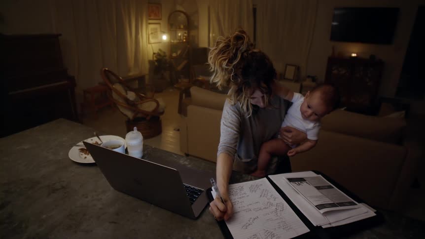 A woman holding a baby scribbles down some notes while cradling a phone receiver and looking at her laptop computer