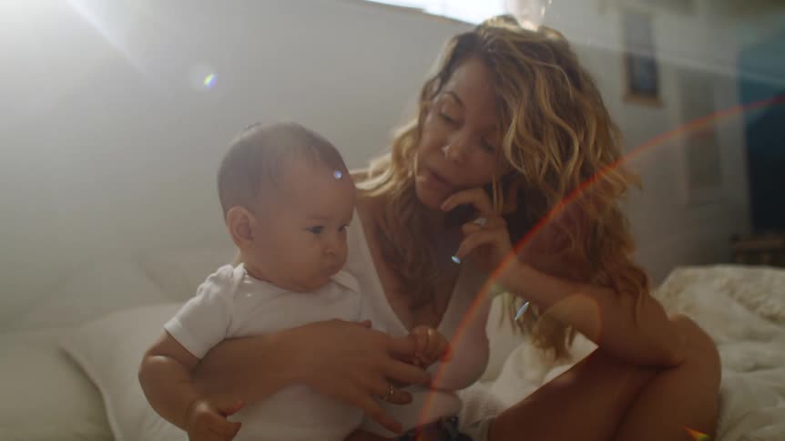 Mother sitting at the bed with her baby son while she talks on the phone during day time