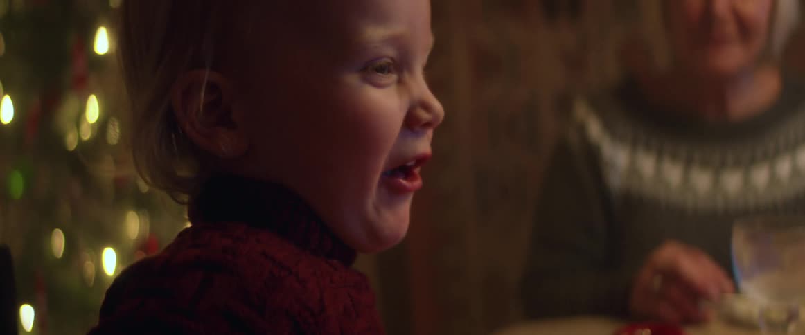 Toddler girl sitting at the table in front of a christmas tree and eating the christmas dinner with her family