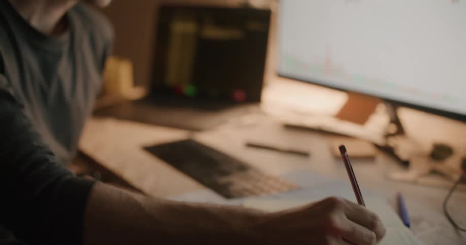 A man sitting at a desk writing notes while looking at his computer monitors