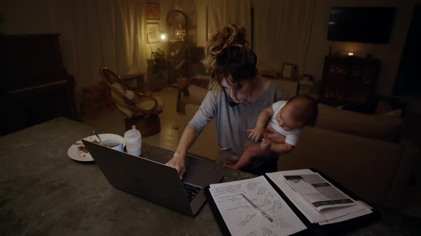 Mother with brown wavy hair and gray sweatshirt holding baby son talks on smartphone and uses laptop in living room at home