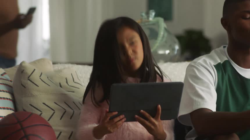 A young girl carrying a tablet sits on a couch next to a teenage boy playing a video game as a girl behind the couch texts and a man walks by with his cell phone