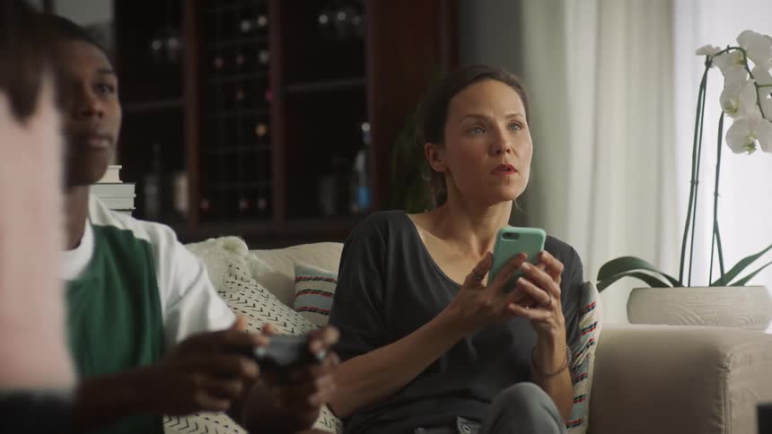 Teenager boy sitting on the couch in the living room playing video game when his mother using her smartphone joins him and then his sister and father