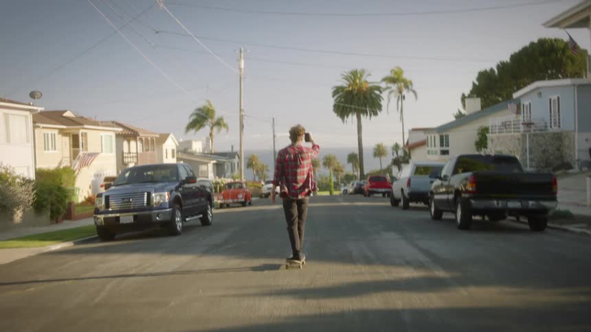 A man skateboards down the center of a neighborhood street holding his phone up filming himself