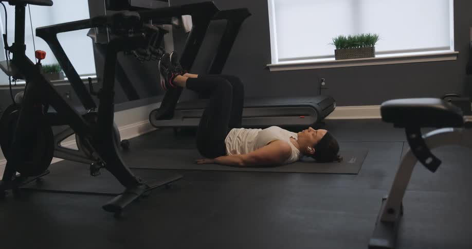 Woman with black headband wireless earphones white shirt black leggings and sneakers does abs workout next to treadmill in the gym