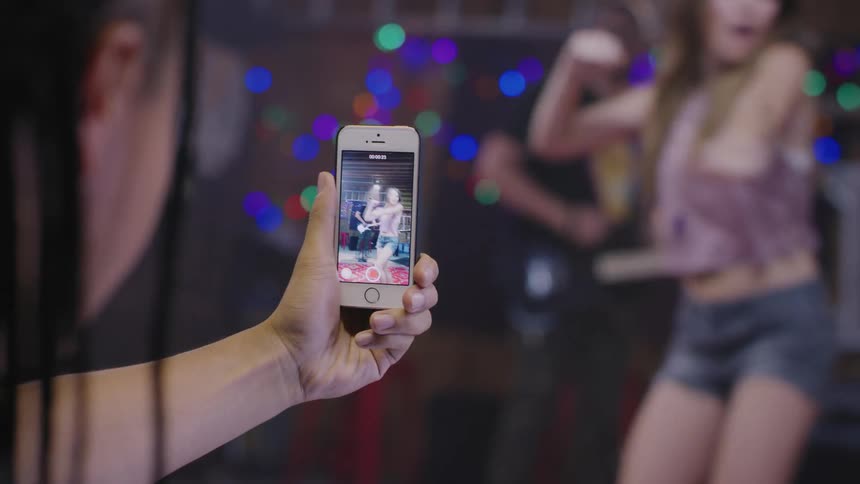 Man using a smartphone to record a woman dancing and a man playing guitar indoors