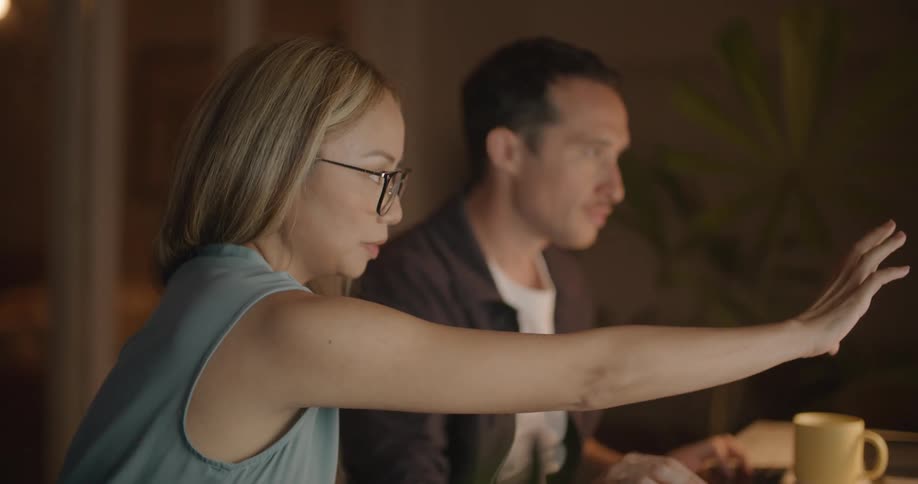 Man with white and black shirt sits at desk using computer and talking to woman with brown hair and blue blouse in the office