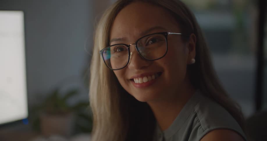 Woman with brown wavy hair glasses earrings and blue blouse talks to coworker in the office