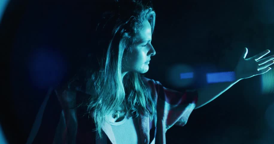 Woman reaches her hand out and examines it against blue spotlight at night