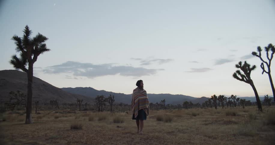 A young woman with a blanket wrapped over her shoulders walks across a field