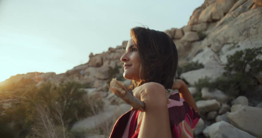 Woman uses her hands to hold a large stick behind her neck while standing near the mountains on a sunny day