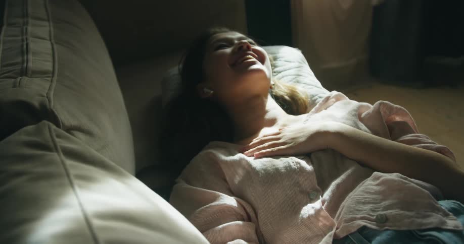 Young woman wearing a wireless earphone lying in the couch with her hand on her chest and laughing