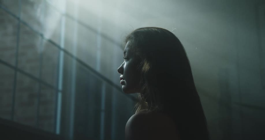 A young woman turns to face the window while standing in a dimly lit room