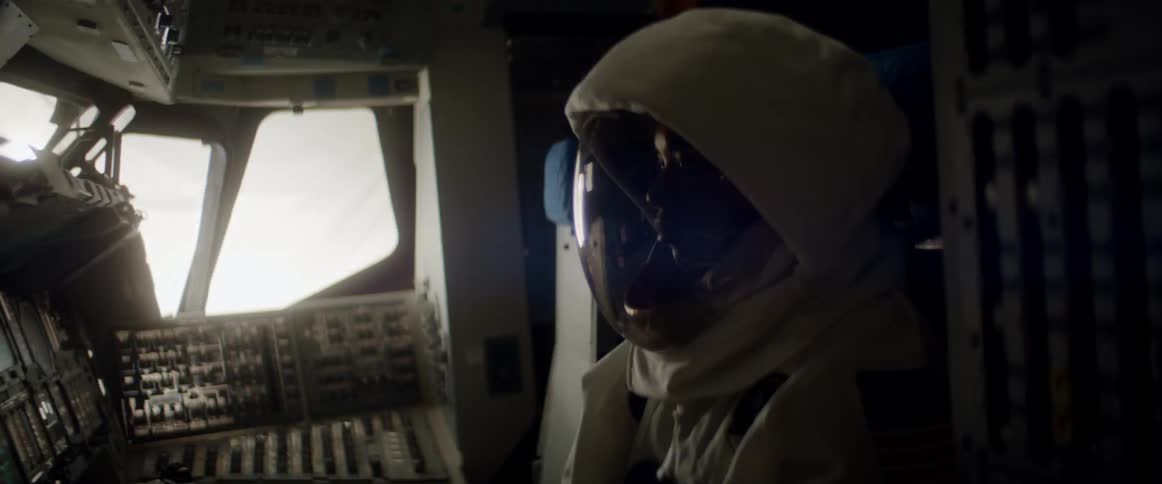 Woman wears spacesuit while sitting in cockpit of space vehicle in front of control panel full of switches