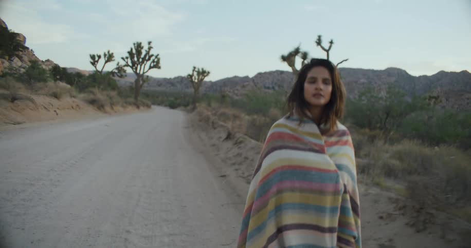 A woman walks along a road through a rugged countryside with a blanket around her shoulders during the day