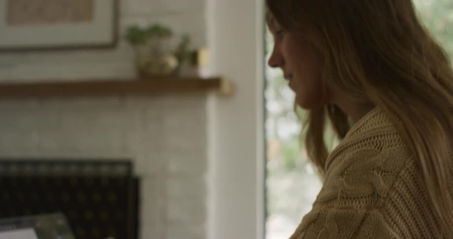 A woman sitting near a fireplace talks and smiles as she types on a laptop