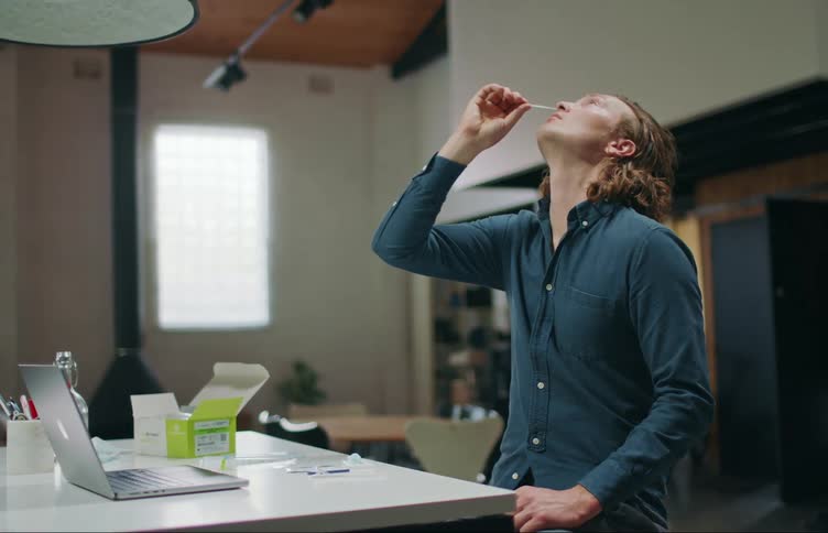 A man sits at a table and leans his head back while he swabs his nasal passage
