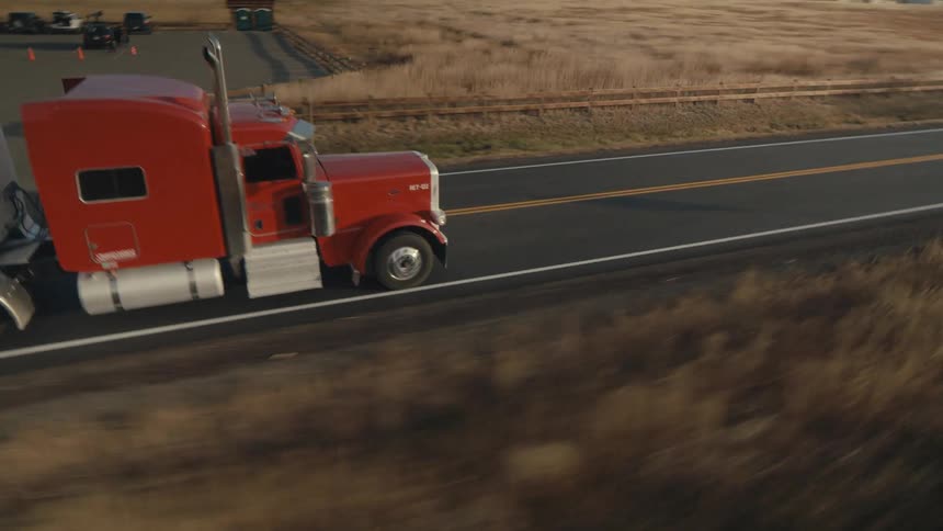 An aerial orbiting view of a tractor trailer traveling on a twolane highway in a rural area in the evening