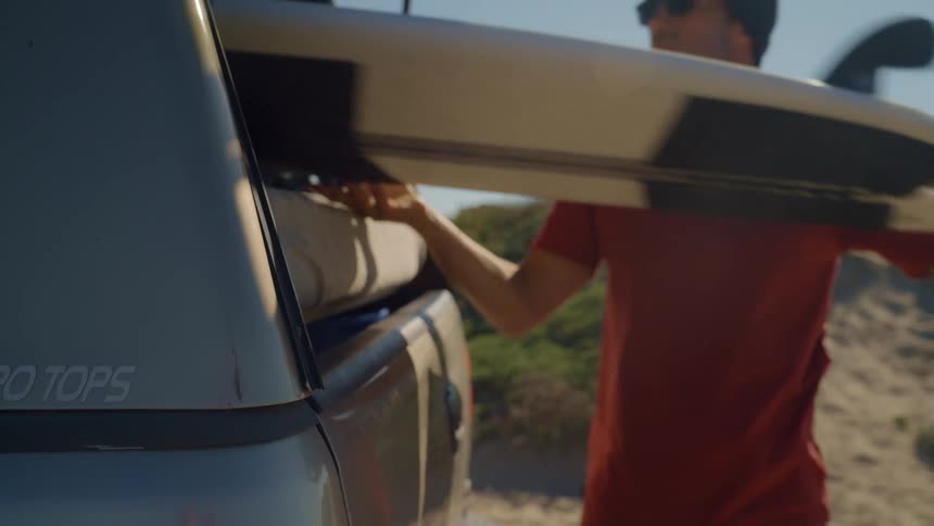 A man opens the back of a vehicle and takes a surfboard out and then a woman walks over and takes a pair of boots out