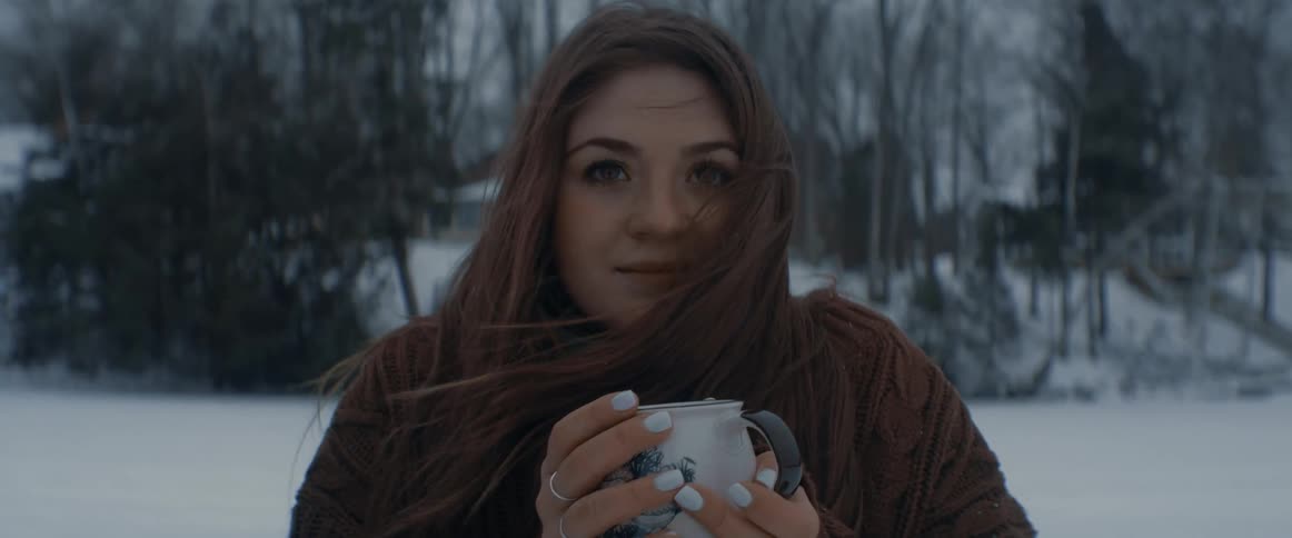 A woman holds a mug in both hands as she stands outside in the snow looking very cold