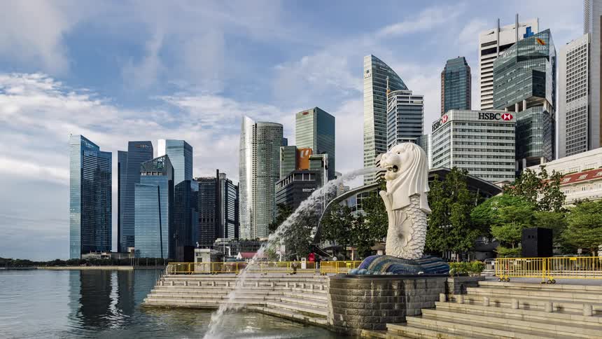 Lion headed fish statue spouts into water as pedestrians move around with city reflecting sky in the background