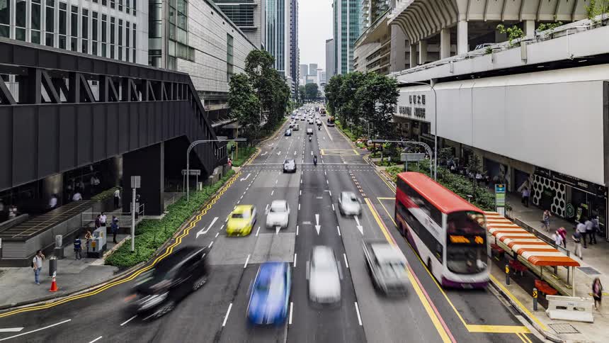 Cars and city buses travel on the city street as people wait at a bus stop and pedestrians walk on the sidewalk and cross the street