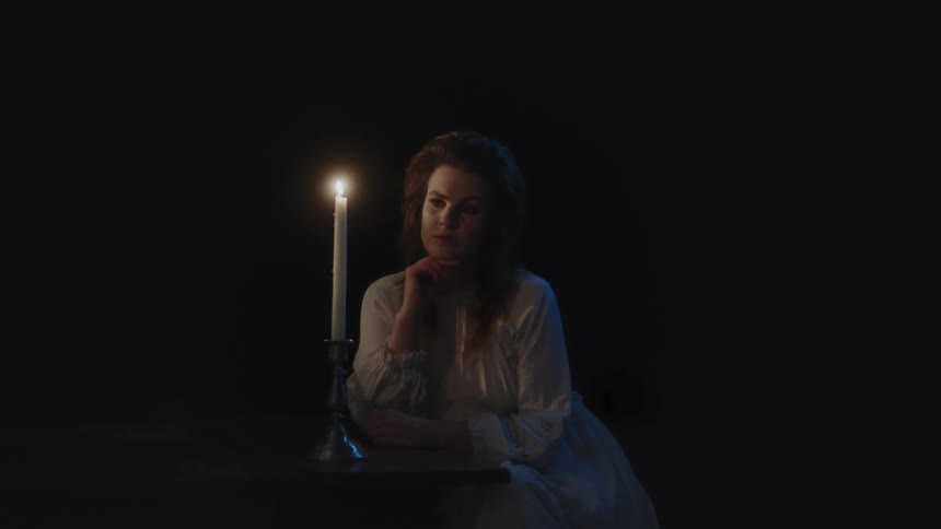 Woman wearing a white gown sitting near a candle over a table inside a dark room at night