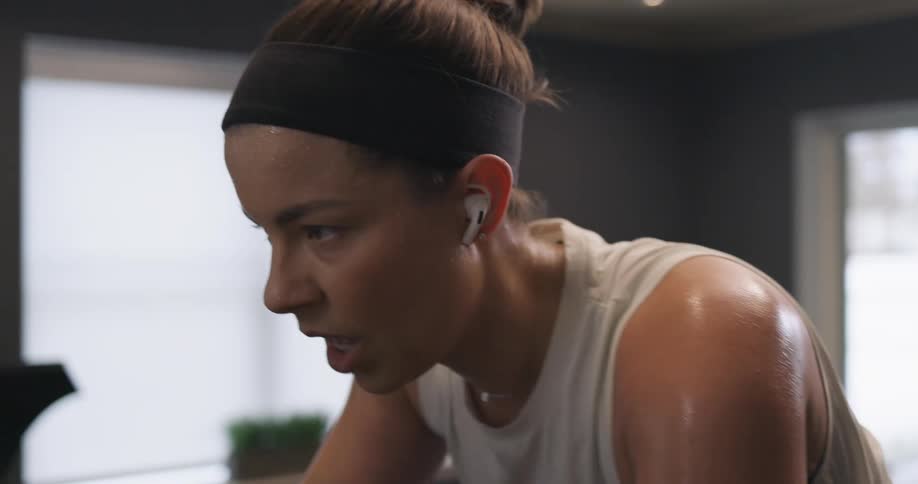 A woman with ear buds in breathes heavily as she rides a stationary bike at home