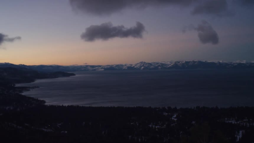 Snowy hill with forest overlooking lake and distant snowy mountain range at dusk