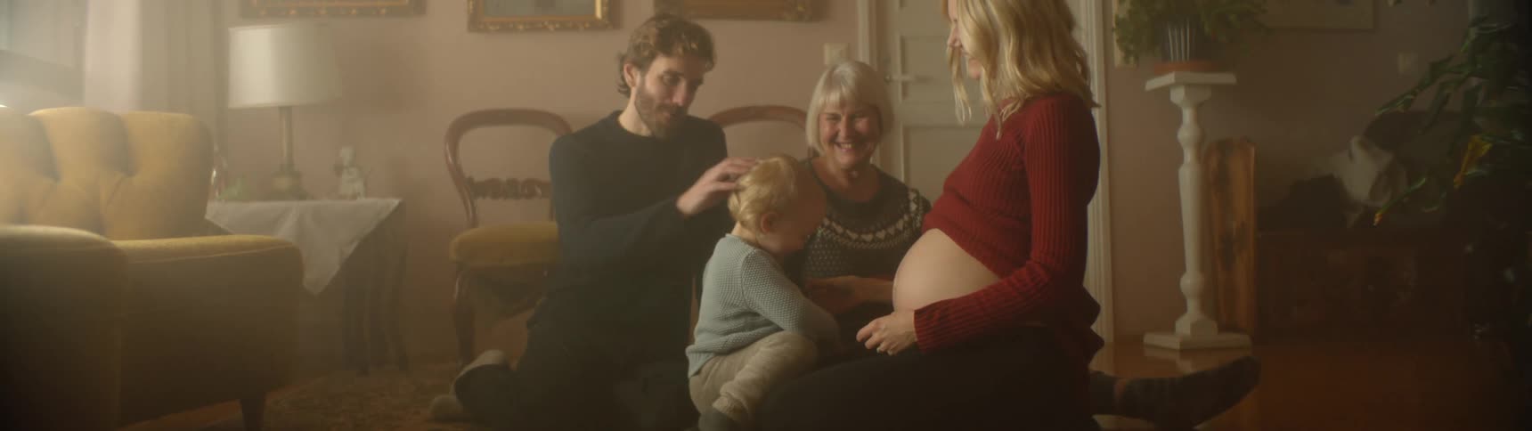 Toddler hugs a pregnant womans bare belly while a man and an older woman watch and smile