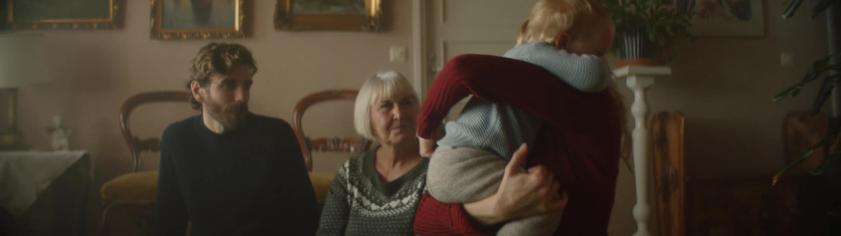 Blonde mother with red sweater holds and cuddles toddler son next to grandmother and father with beard and black shirt