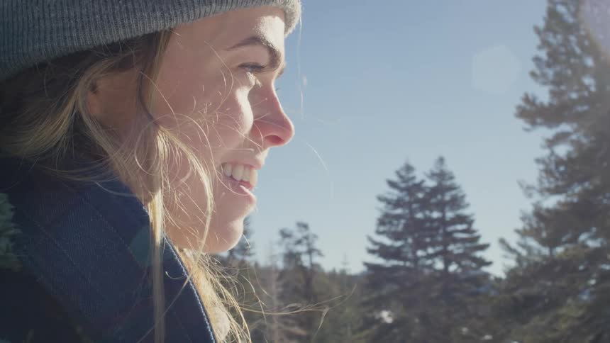 Blonde woman with gray wooly hat and green jacket looks around smiling on snowy hill next to trees as the sun shines on face