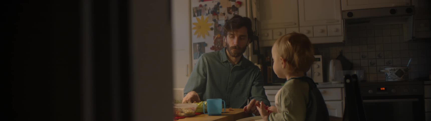 Father with beard and blue shirt sits at table with son eating food in the kitchen