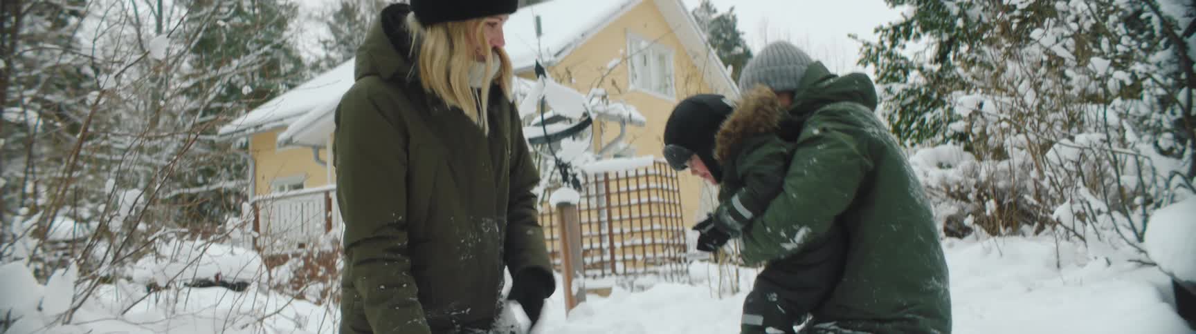 Family wearing thick winter clothes playing with snow on the ground near a house at winter