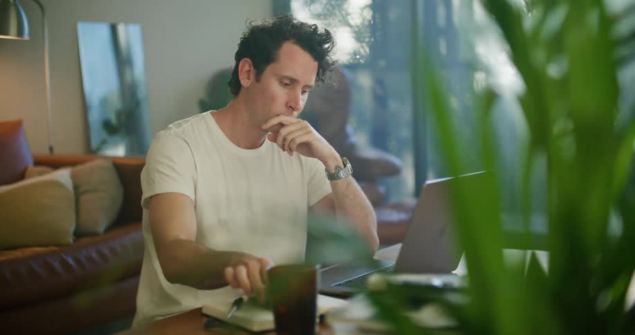 A man sits at a table with his laptop while checking his cell phone for a message