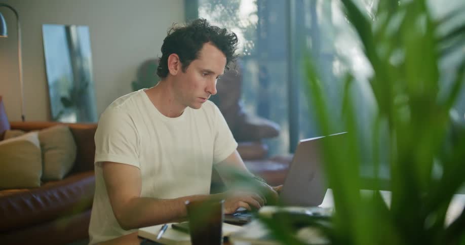 Man in a white short sleeved shirt and wearing a wristwatch types on a laptop on a table with a pen laying nearby