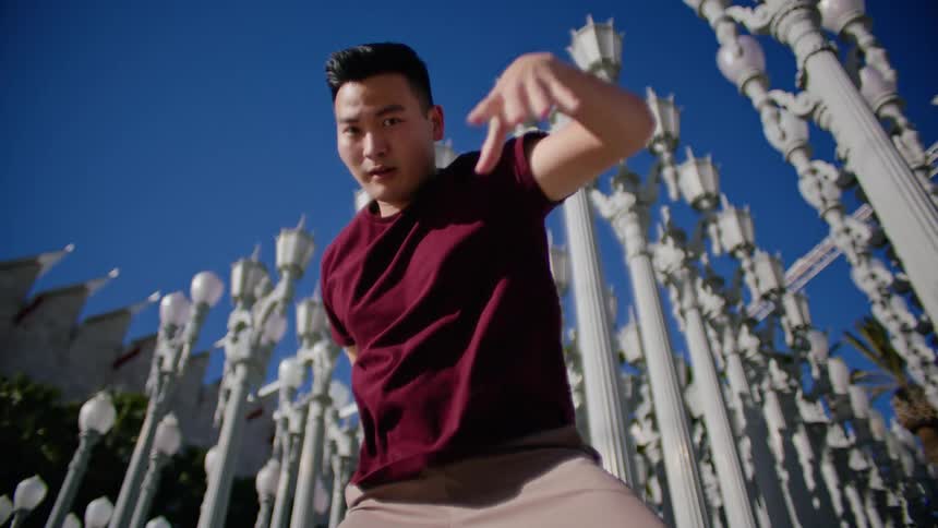 An asian male looks directly at the camera as he performs a dance outdoors near many lampposts