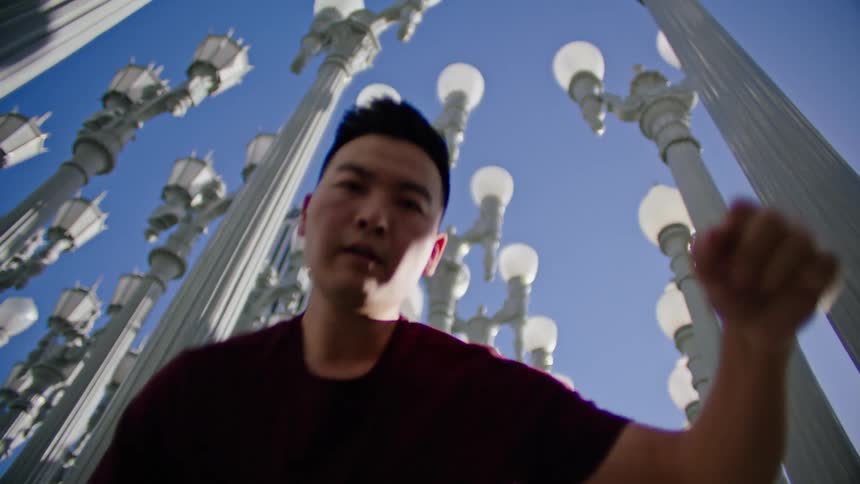An asian male looks directly at the camera as he performs a dance in front of some lamp posts