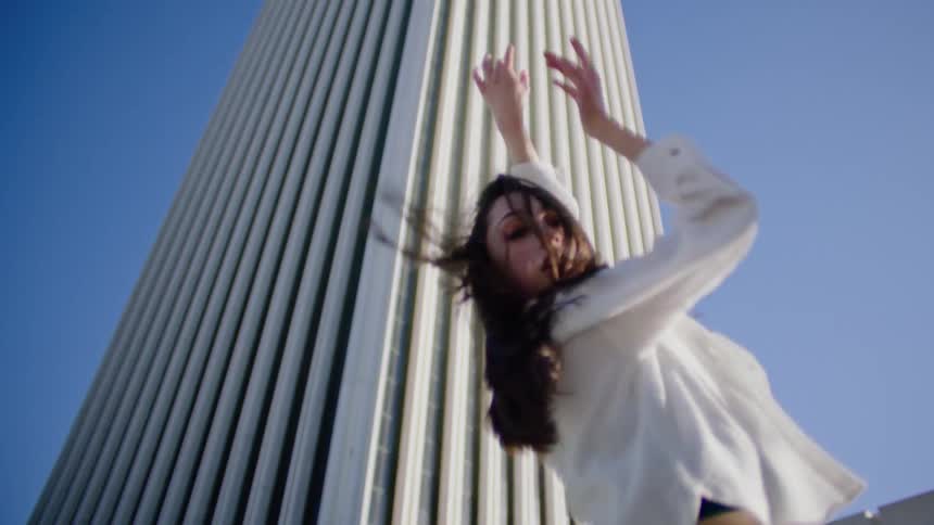 A young woman dances outdoors in front of a towering skyscraper during the day