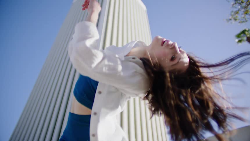 A female dancer performs a series of dance moves outside in front of a tall building on a very windy day
