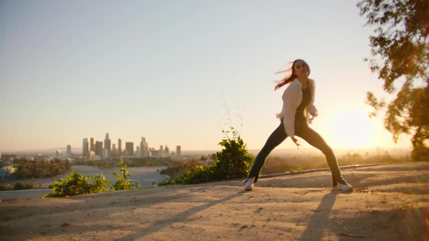 Female dancer with white shirt and teal top and pants dances against city skyline at sunset