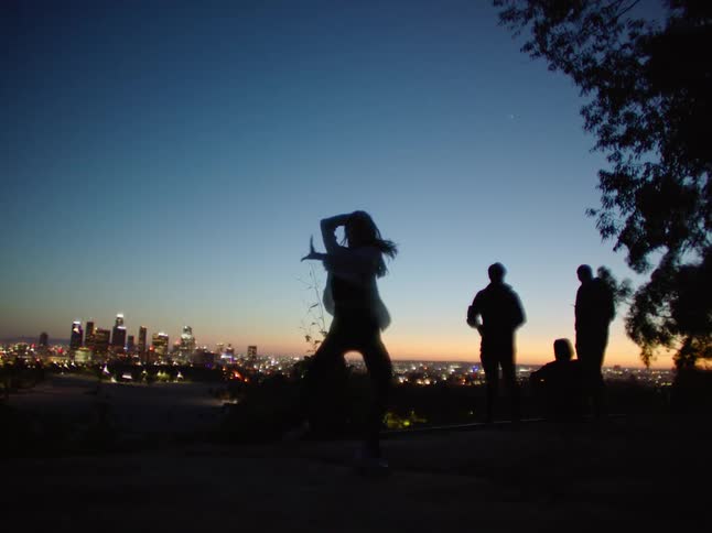 Silhouette of a young woman energetically dancing in front of friends next to the city skyline at sunset