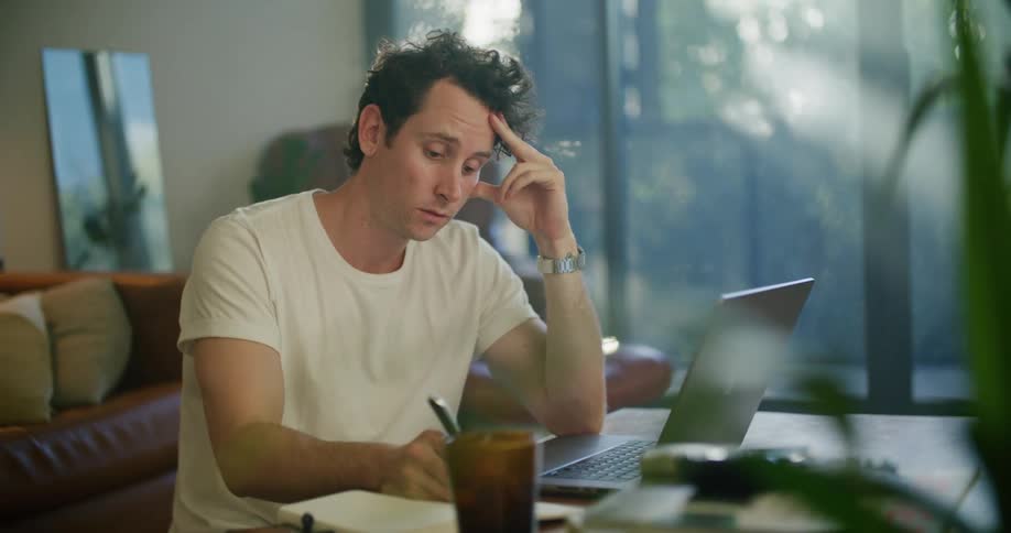 Man with white t shirt works on laptop and writing in notebookat coffee table next to brown couch in living room at home